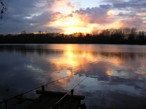 The sun sets on a winter's day at Kingsmead One fishing lake, two rods pointing out into the lake with the sun dipping behind the bare tree line on the far bank.